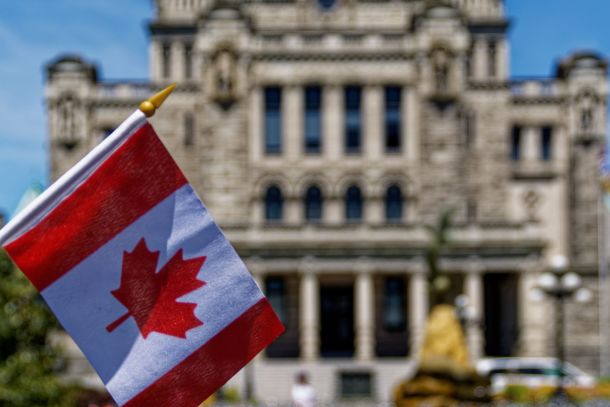 A Canadian flag in focus with a blurred background of a historic stone building and trees under a clear sky.